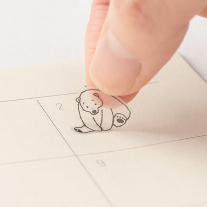 Small bear-shaped sticker on a planner page with a hand pressing it down.