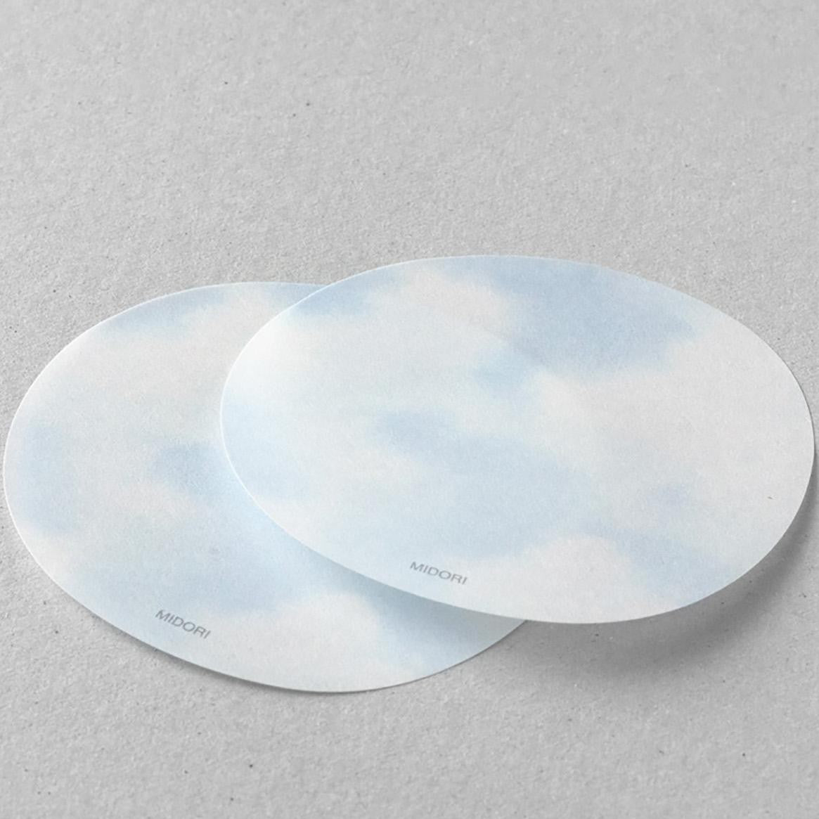 A hand peeling a translucent sticky note from a circular sky-themed pad with cloud design
