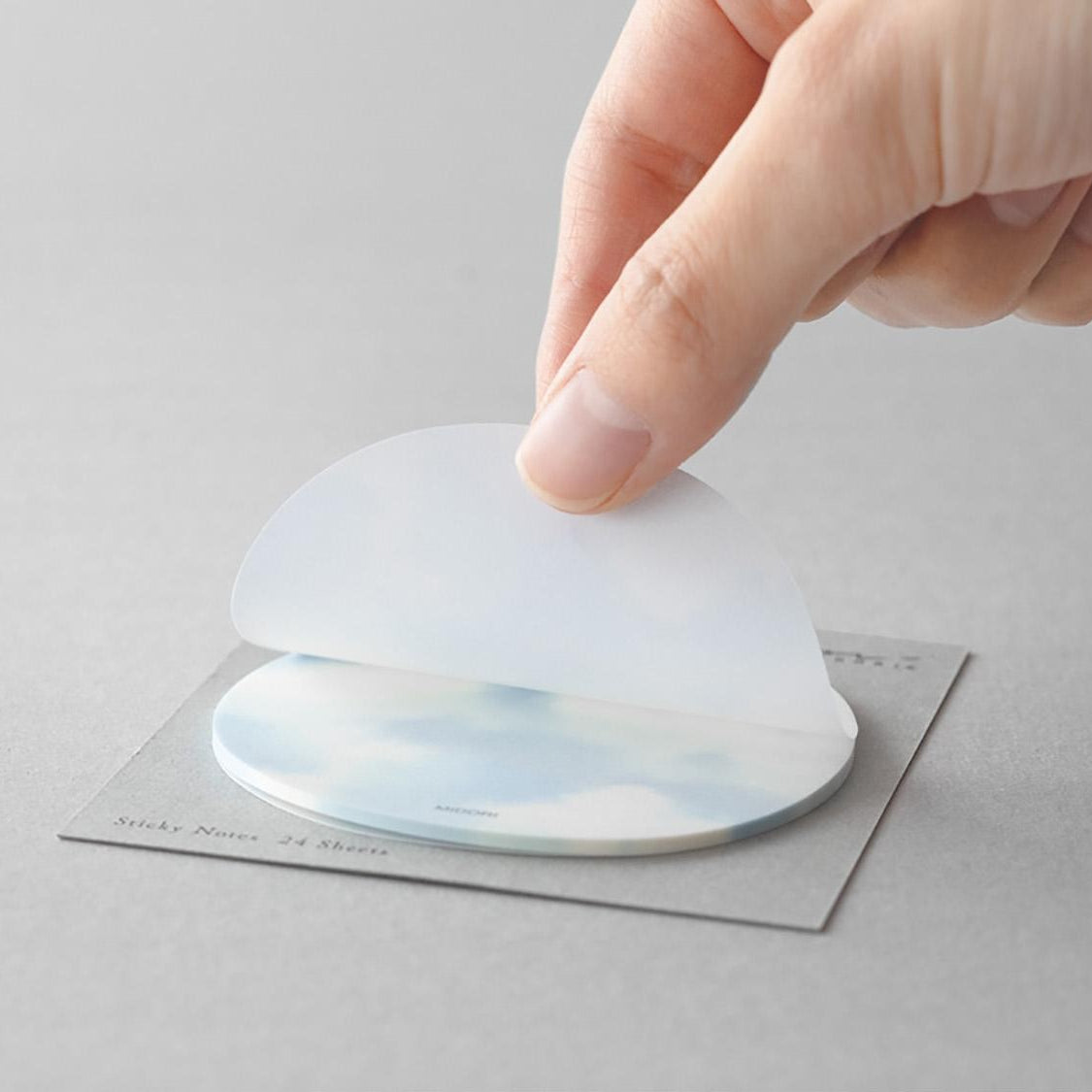 A hand peeling a translucent sticky note from a circular sky-themed pad with cloud design
