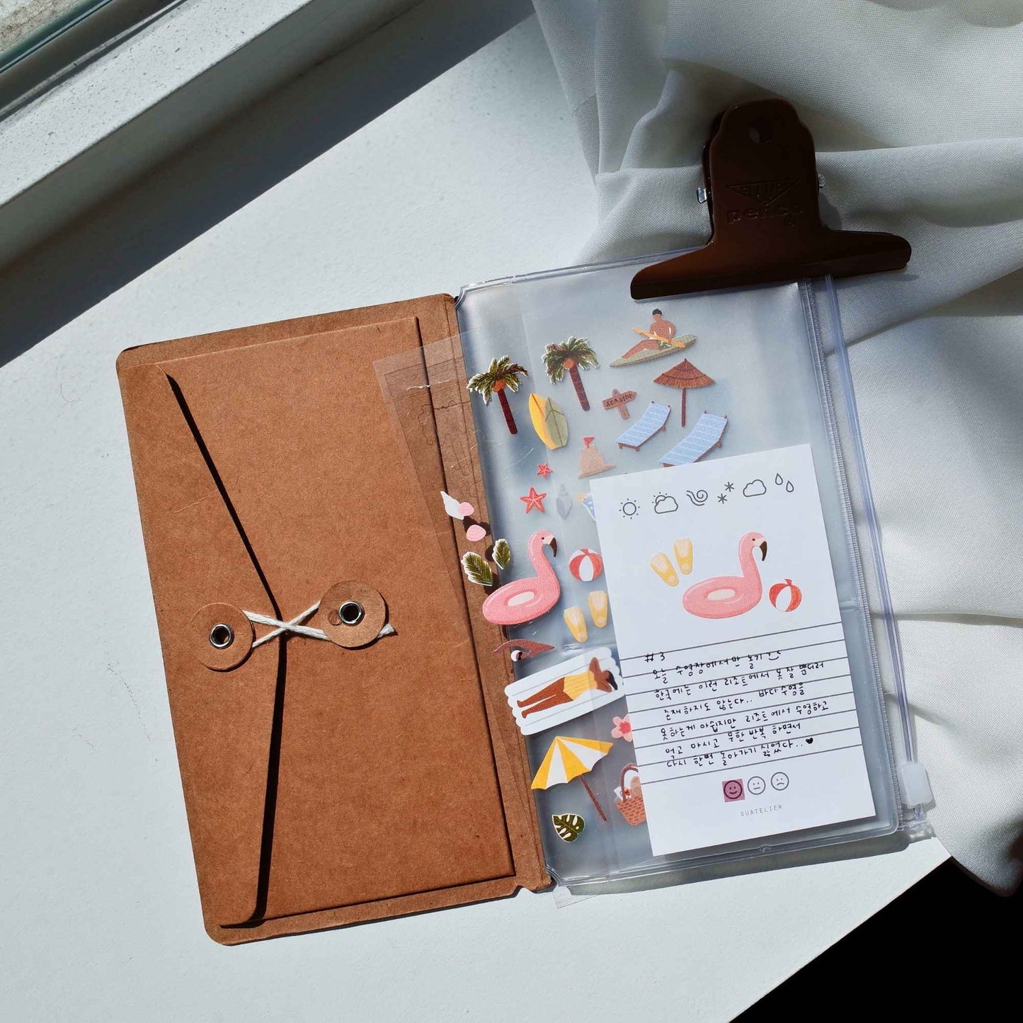 A clear planner pocket with a brown leather tab and a brown envelope-style cover, shown open on a white surface with sunlight and white fabric nearby. The pocket contains decorative stickers, a pink flamingo note card, and small accessories.